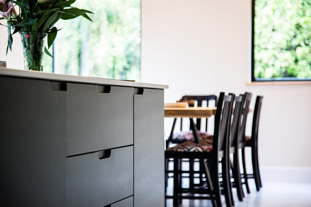A modern kitchen with a dark island, vase of flowers, and a dining table with black chairs near large windows showing greenery outside.