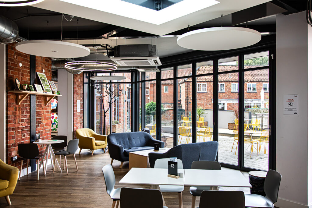 Modern café interior with lounge chairs, tables, and large windows overlooking an outdoor patio with yellow chairs and tables. Exposed brick walls and contemporary lighting fittings are visible.