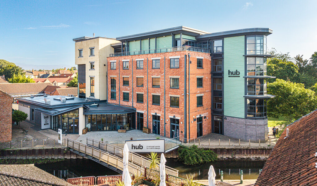 Modern multi-storey building labelled “hub” by a canal, featuring brick and glass exteriors, a footbridge, and surrounding greenery under a blue sky.