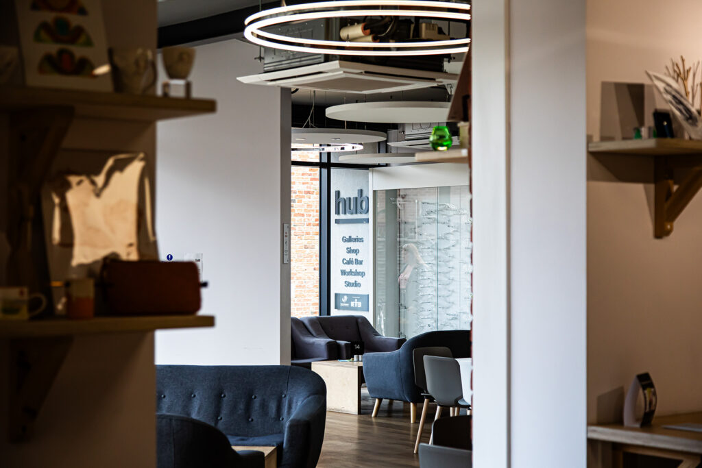 A modern café interior with grey sofas, round ceiling lights, and a sign reading “hub: Gallery, Shop, Café Bar, Workshop, Studio” visible through a doorway.