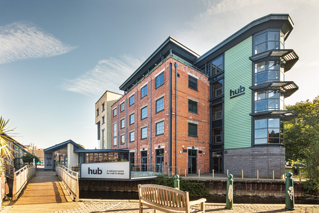 Modern multi-storey building labelled hub, with a mix of brick and green cladding, situated by a canal with a bench and trees in the foreground.