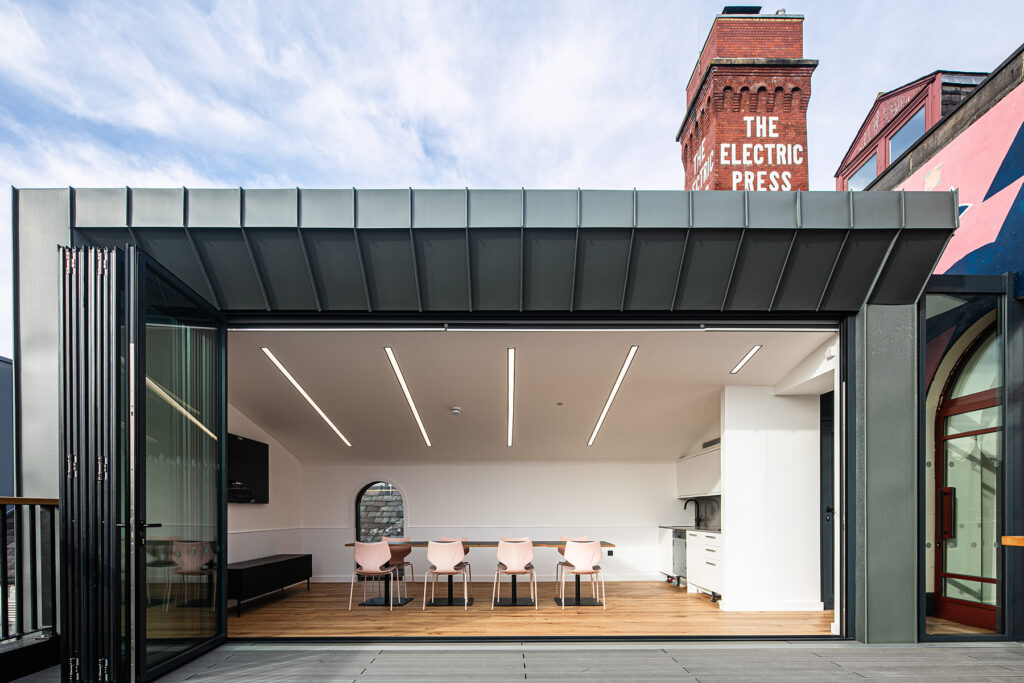 Modern meeting room with pink chairs and a kitchenette, open to a terrace; the Electric Press brick tower visible in the background.