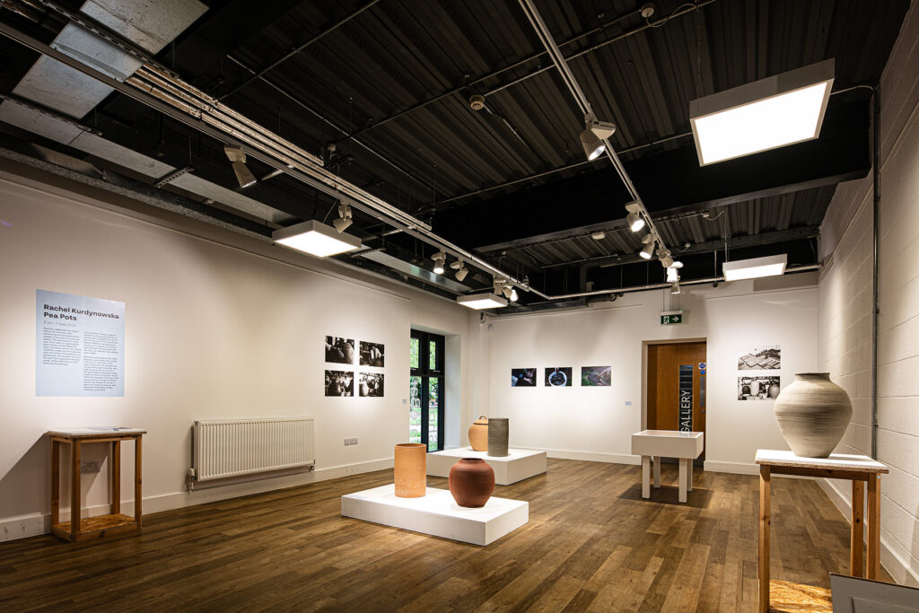 A modern art gallery room with ceramic pots displayed on white plinths, framed photographs on the walls, and a wooden floor under bright ceiling lights.