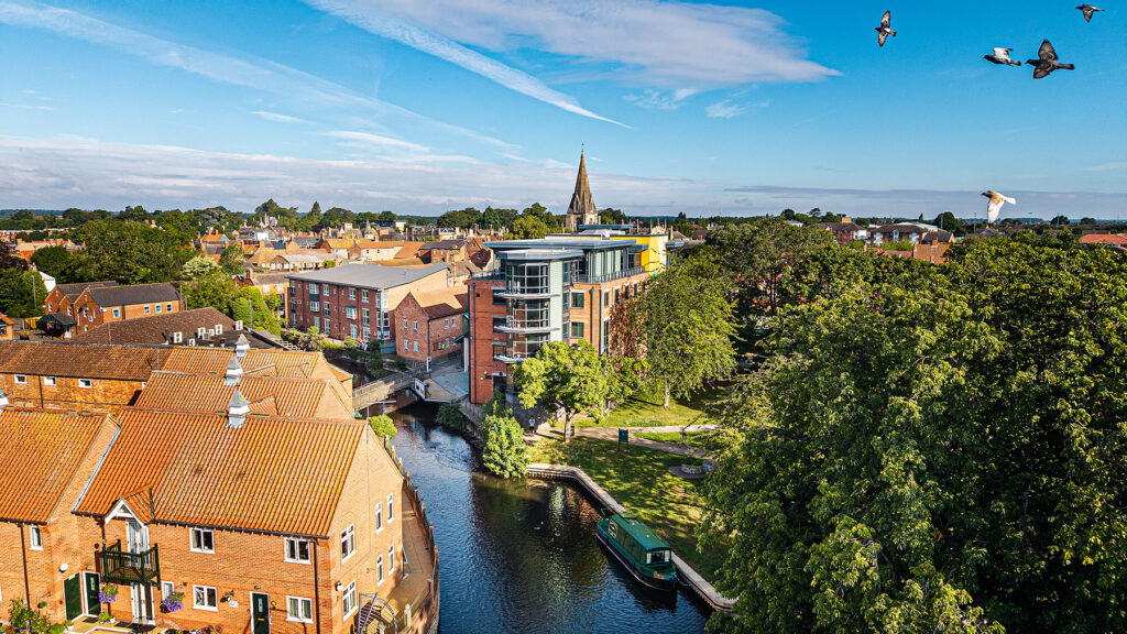 Aerial view of a riverside town with red-roofed buildings, modern architecture, a church spire, green trees, and several birds flying in a blue sky.