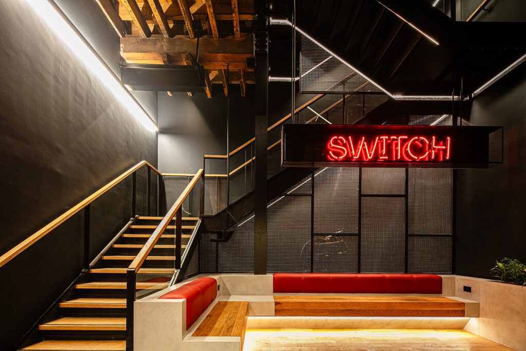 A modern interior with black metal stairs, wood accents, red cushioned seating, and a red neon SWITCH sign on the wall.