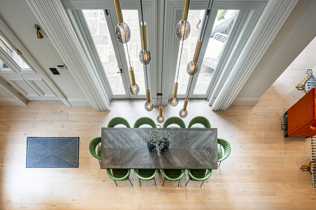 Overhead view of a rectangular dining table with eight green chairs, a vase with branches in the centre, and pendant lights hanging above in a bright room with wood flooring. 