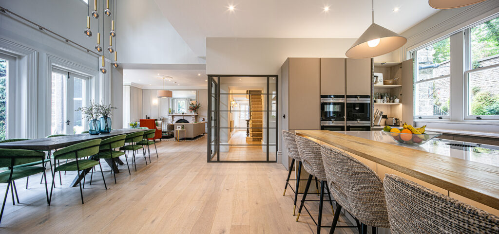 Modern open-plan kitchen and dining area with light wood floors, large windows, a central island with bar stools, and a dining table with green chairs.