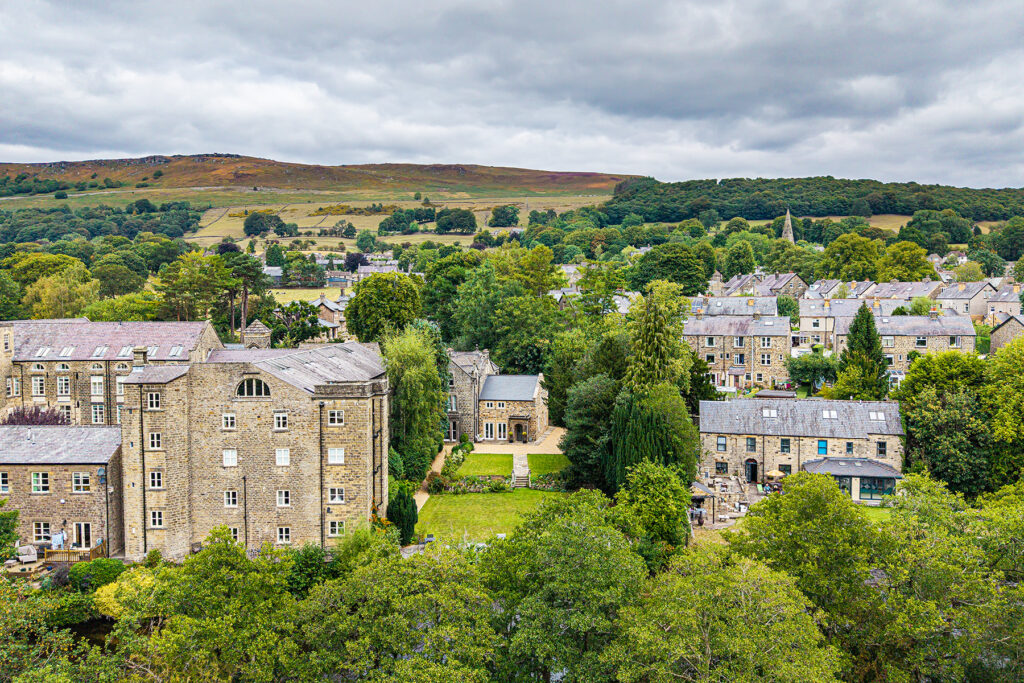 Aerial view of stone buildings surrounded by trees in a small village, with hills and cloudy sky in the background.