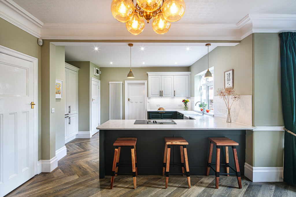 Modern kitchen with green walls, wood herringbone flooring, a white island worktop, three wooden bar stools, and pendant lighting above the island.