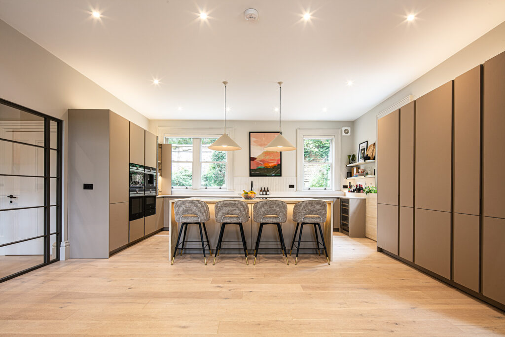 Modern kitchen with neutral cupboards, an island with four bar stools, pendant lights, built-in ovens, and large windows letting in natural light. 