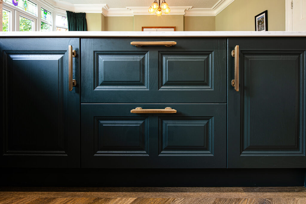 Navy blue kitchen cupboards with gold handles and a white worktop, set against a background of wooden flooring and light-coloured walls. Photograph by Dug Wilders.