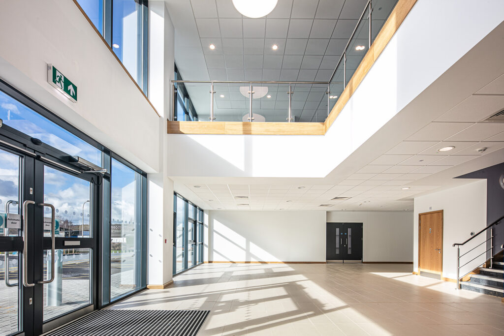 A spacious, modern building lobby with large windows, glass doors, stairs, and a mezzanine level with glass balustrade, filled with natural light.