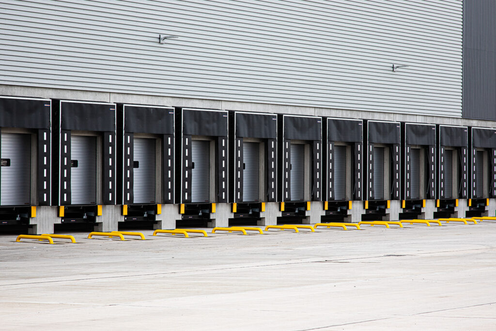 A row of closed loading bay doors with black bumpers and yellow barriers in front, attached to a modern industrial building.