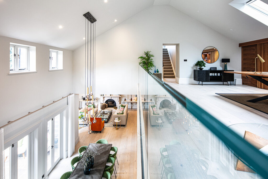 Modern open-plan home interior with a mezzanine level, glass balustrade, dining area, lounge, and staircase leading to an upper floor. Natural light fills the space through multiple windows. 