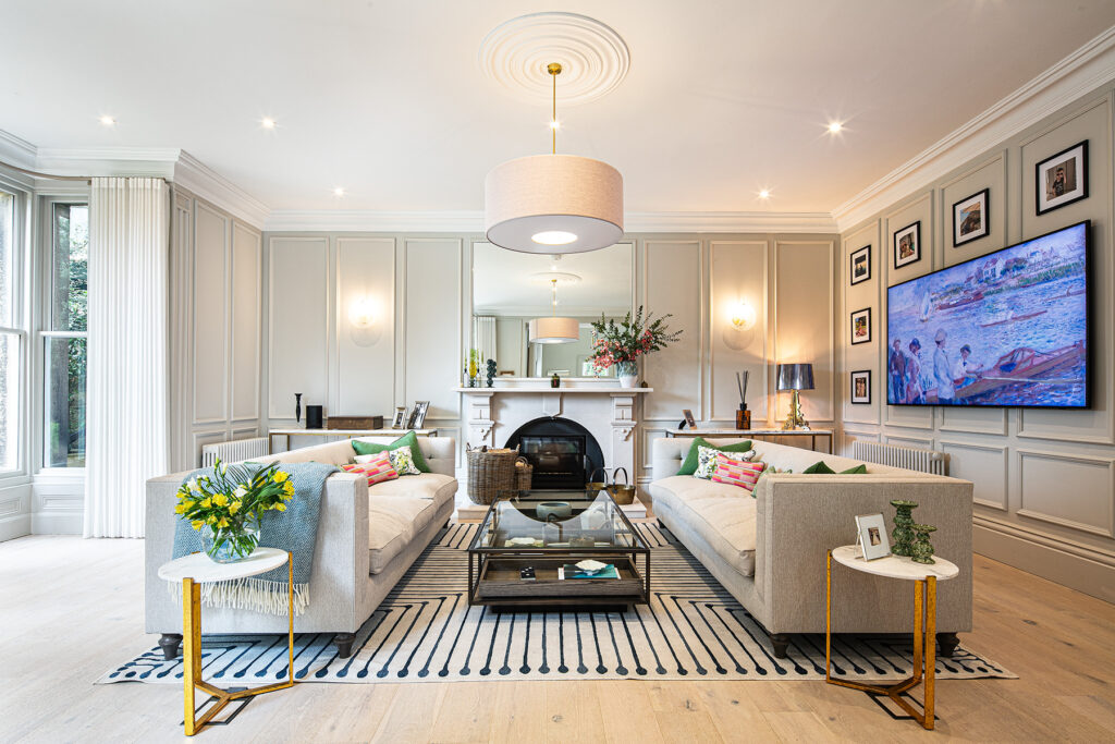 Elegant sitting room with two beige sofas, striped rug, glass coffee table, wall-mounted TV, fireplace with mirror, and decorative lighting. Large windows let in natural light.