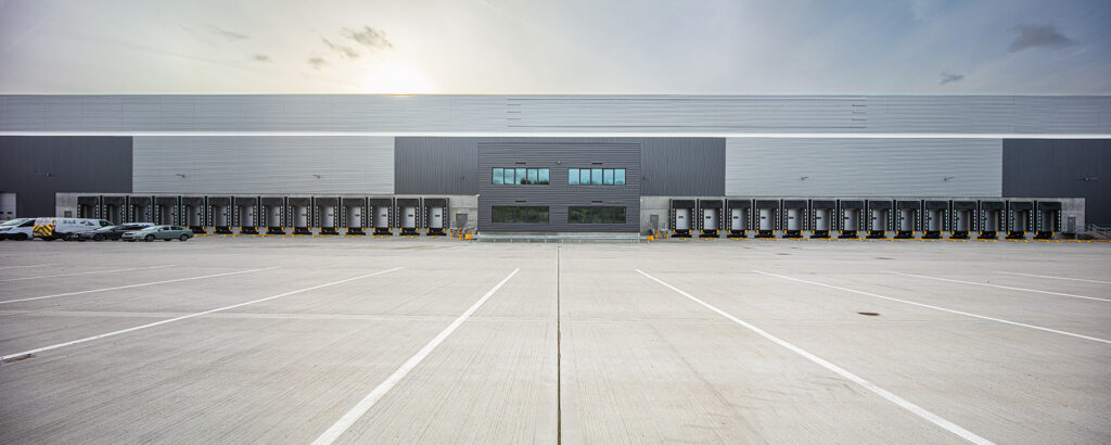 Large modern warehouse with multiple loading bays, a central entrance, and a mostly empty car park under a partly cloudy sky.