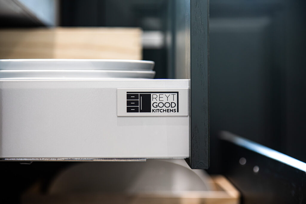 A close-up of an open kitchen drawer with a Reyt Good Kitchens label, showing stacked white plates inside.