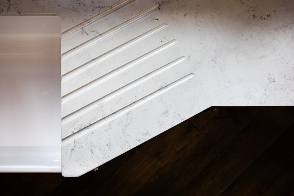 White worktop with four parallel grooves next to a white sink, with a marble-like pattern and dark wood floor below.