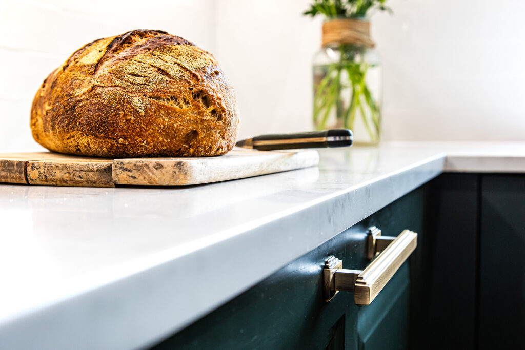 A loaf of bread sits on a wooden chopping board with a bread knife on a white kitchen worktop; a jar with greenery is in the background.