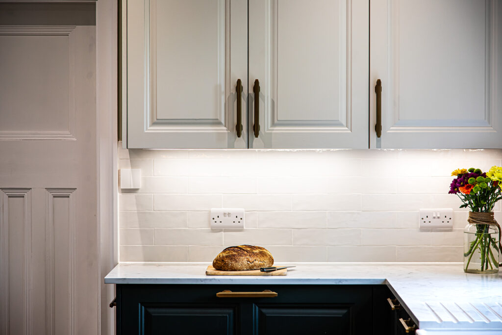 A loaf of bread and a knife on a marble worktop beneath white cupboards, with a vase of flowers to the right in a modern kitchen.