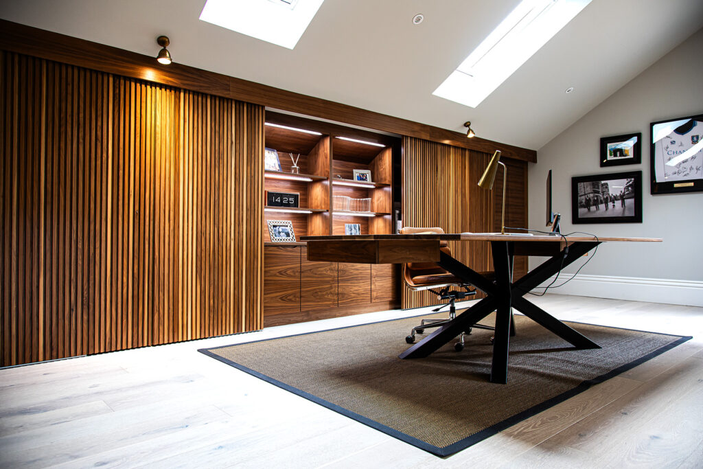 Modern home office with wood panelled walls, built-in shelves, a large desk, swivel chair, rug, skylights, and framed art on the wall.