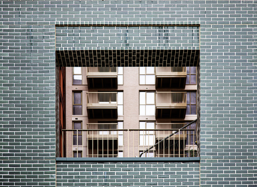 A brick wall with a large square opening frames a view of a modern block of flats with balconies.