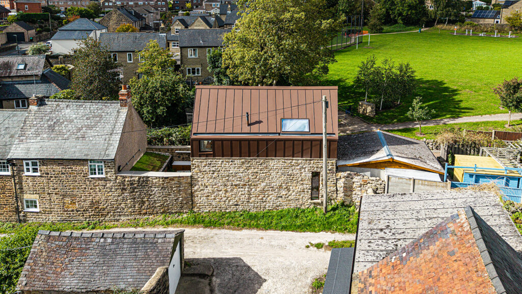 A modern house with a brown metal roof and stone walls sits between older stone buildings, next to a grassy field and trees in a residential area.