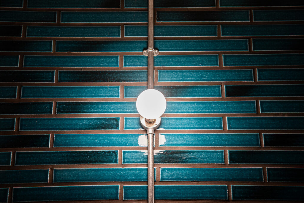 A round white light bulb is mounted on a wall with horizontal rectangular teal tiles and visible metal piping.