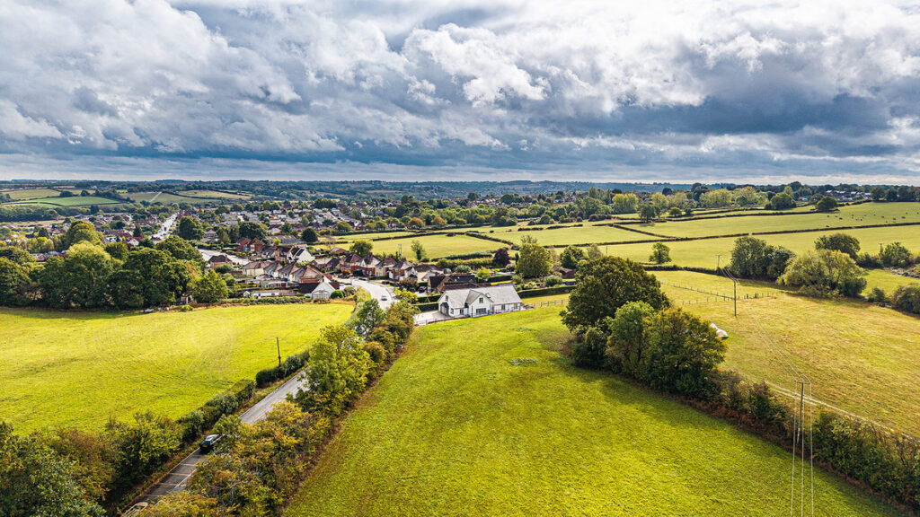 Aerial view of a rural landscape with green fields, scattered trees, a small village, and cloudy skies.