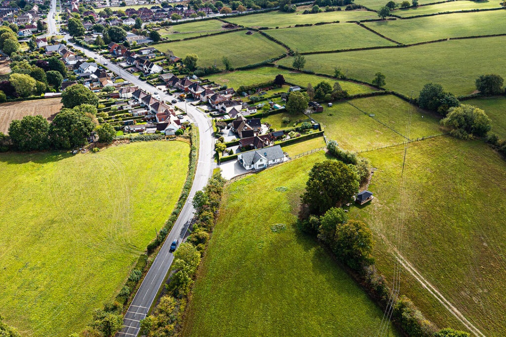 Aerial view of a rural area with houses, a road, open green fields, and scattered trees on a sunny day.