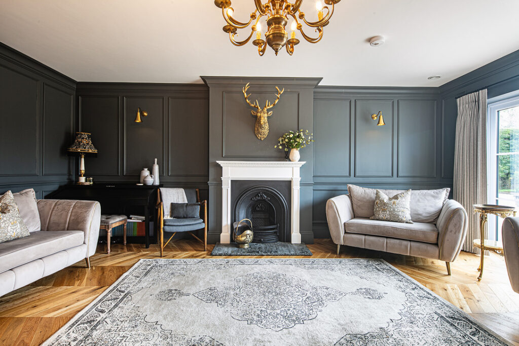 Elegant sitting room with dark panelled walls, a white fireplace, gold chandelier, plush seating, and a decorative stag’s head above the mantelpiece. Light flows in from a window on the right.