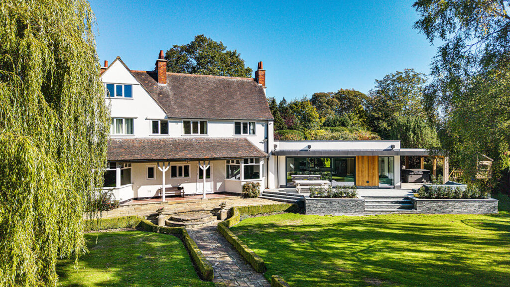 Large two-storey house with white exterior, dark roof, and modern extension, surrounded by a landscaped garden with a stone path and mature trees.