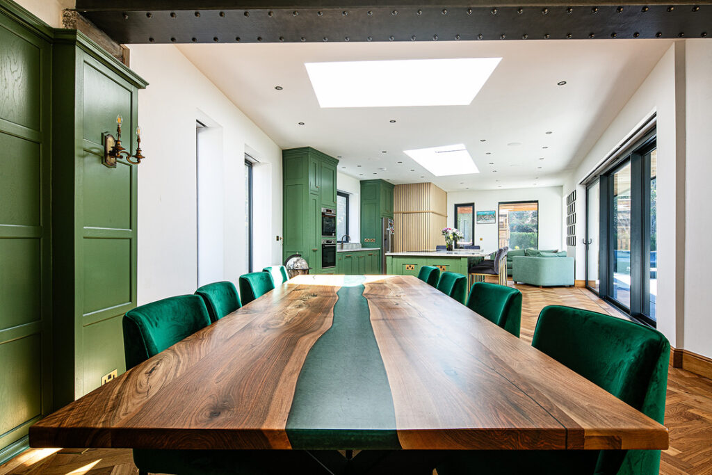 Modern dining room with a long wooden table, green upholstered chairs, green cabinetry, large windows, and skylights providing ample natural light.