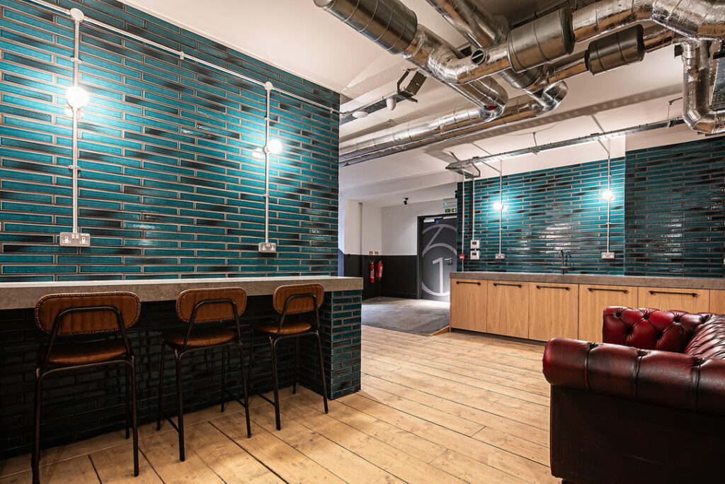 Modern interior space with teal brick walls, exposed ducts, wooden floor, bar-height counter with chairs, wooden cabinets, and a red leather sofa. Brightly lit with multiple wall lights.