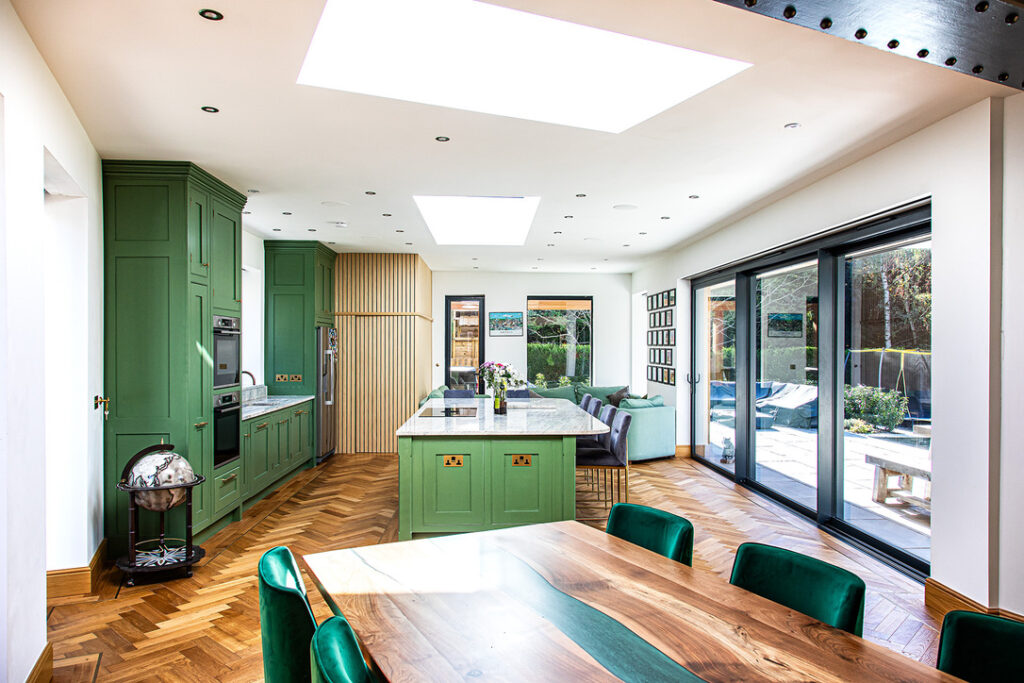 Modern kitchen with green cupboards, a large island, skylights, herringbone wooden flooring, and glass doors opening to a patio. A wooden dining table with green chairs is in the foreground.