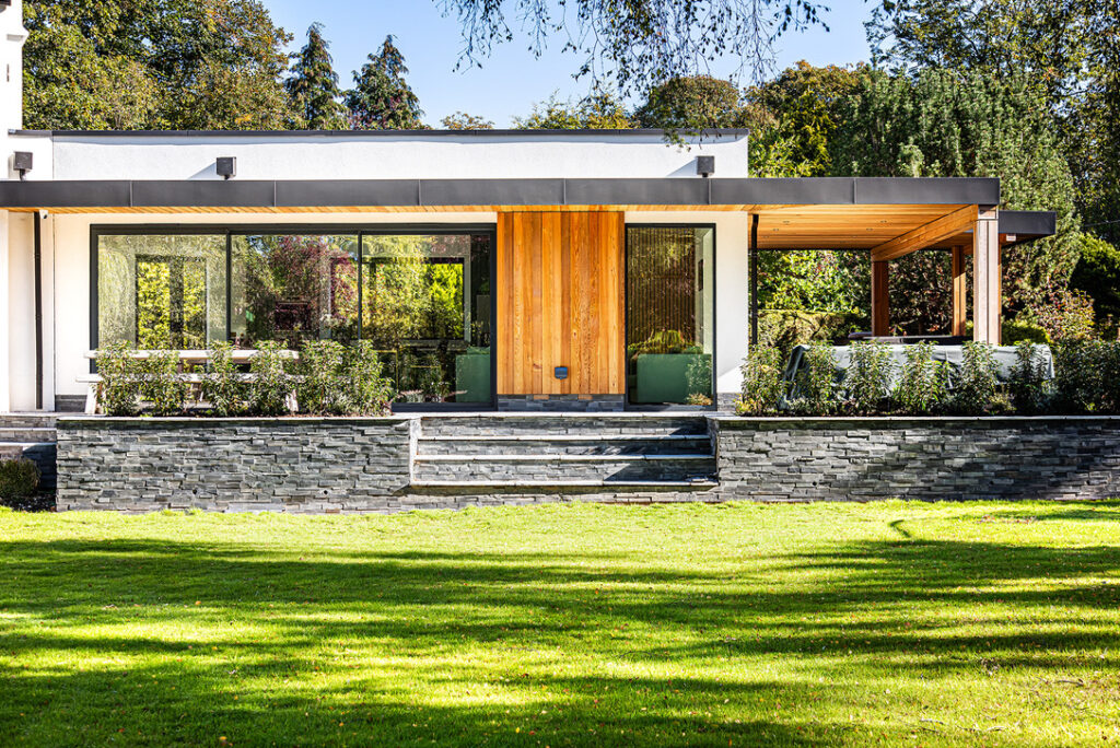 Modern single-storey house with large windows, wooden accents, and a stone porch, surrounded by greenery and a well-kept lawn.