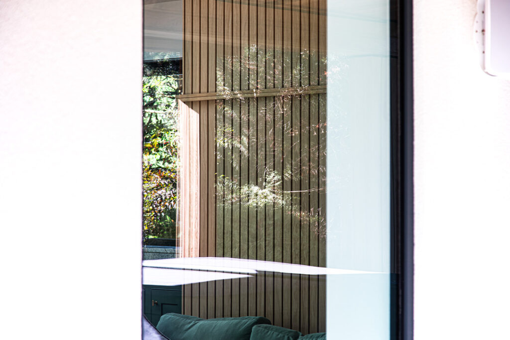 Reflection of trees and vertical wooden slats seen in a large glass window, with part of a green sofa visible through the glass.