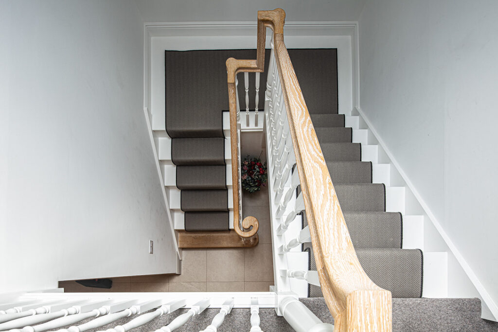Top-down view of a staircase with grey carpet runners, white railings, wooden banister, and a small floral arrangement at the bottom on a tiled floor.