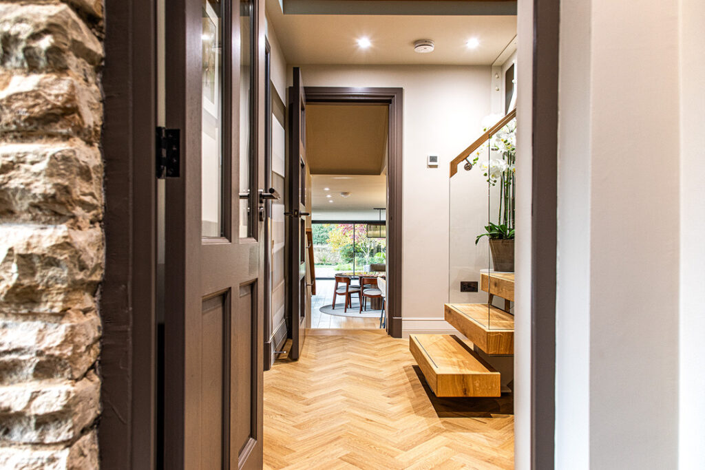 A bright hallway with wood herringbone flooring, floating wooden stairs with glass balustrade, and a view into a dining area with large windows.