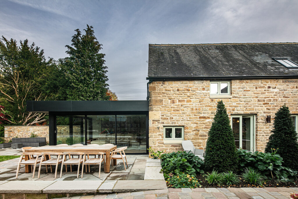 A modern extension with large glass windows connects to a traditional stone house, with an outdoor dining area and landscaped garden in the foreground.