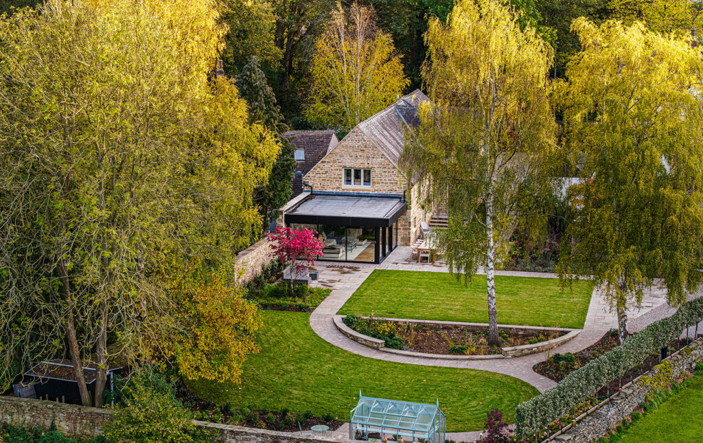 A stone house with a modern extension sits in a landscaped garden with a lawn, curved paths, trees, and a small glasshouse, surrounded by dense greenery.