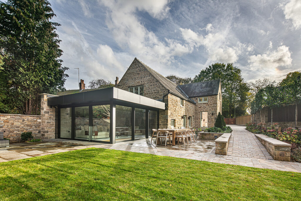 Modern glass extension attached to a traditional stone house, with a paved patio, outdoor dining area, lawn, and landscaped garden path. 