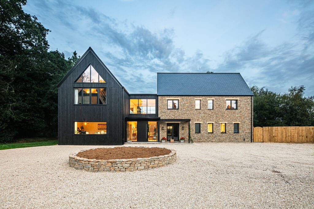A modern two-storey house with a mix of black timber and stone exterior, large windows, and a circular stone planter in a gravel drive.