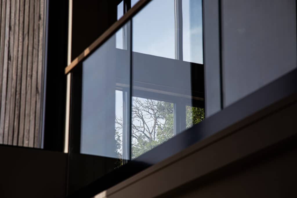Close-up of a modern glass balcony balustrade with reflections and a view of trees and sky through windows in the background.