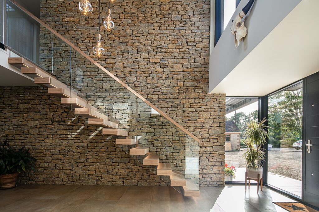 Modern interior entryway with floating wooden staircase, glass balustrade, exposed stone wall, large windows, and hanging pendant lights.