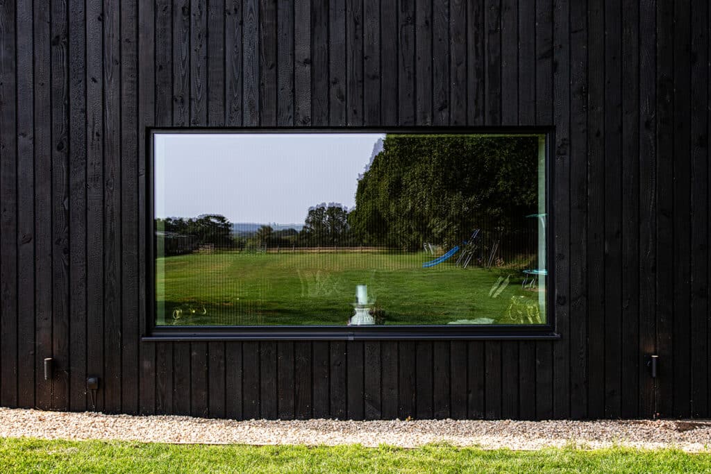 Large rectangular window on a dark wooden exterior wall reflects a green lawn, trees, and playground equipment under a clear sky.