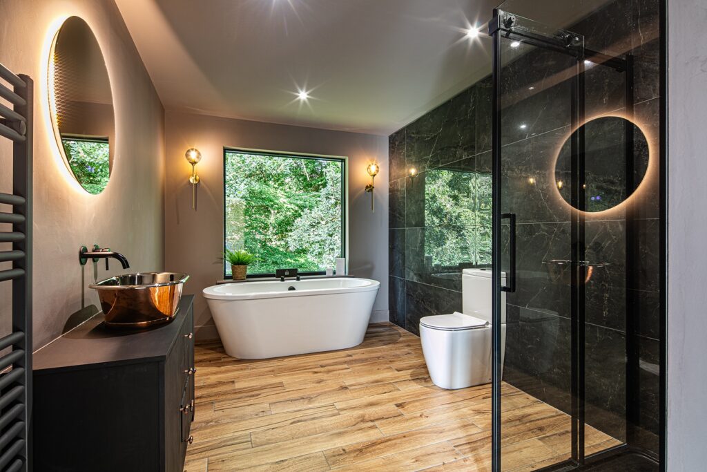 Modern bathroom with a freestanding bath, glass shower, black wall tiles, wooden floor, round mirrors with lights, and a large window overlooking greenery.