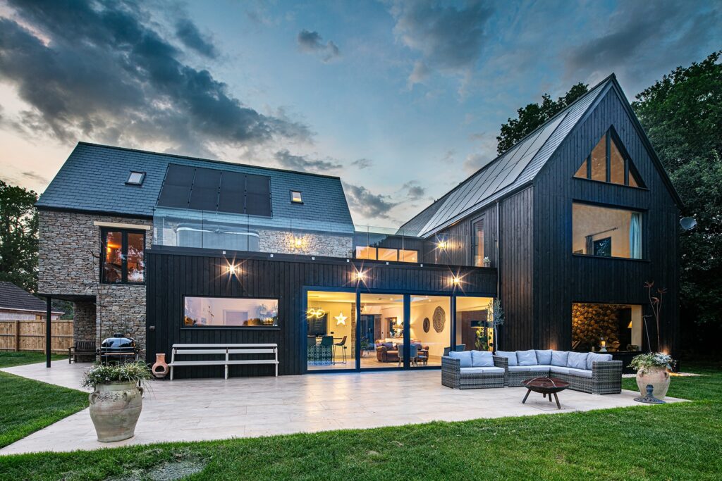 Modern house with a mix of stone and dark wood exterior, large windows, outdoor seating area, patio, and lawn, photographed at dusk.
