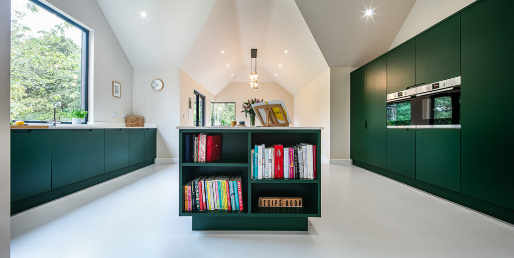 Modern kitchen with green cupboards, a central island featuring open shelves with cookery books, built-in ovens, large windows, and abundant natural light.