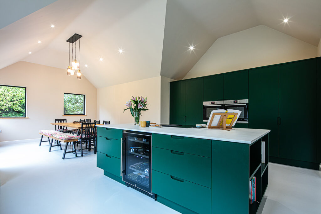 Modern kitchen with green cupboards, white worktops, a central island with built-in wine cooler, dining table with benches, pendant lights, and large windows providing natural light.